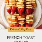 A plate with french toast kebabs—skewers of French toast cubes and strawberries arranged alternately—displayed above text reading Valentines Day Food: French Toast Kebabs. The image has a soft, warm tone.
