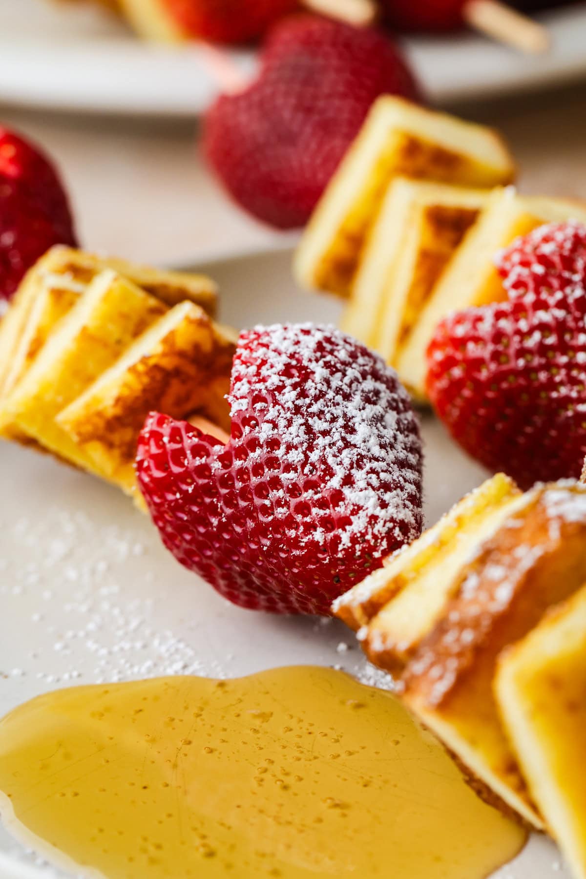 Close-up of french toast kebabs with fresh strawberries and waffle pieces, dusted with powdered sugar, served on a plate with a drizzle of syrup.