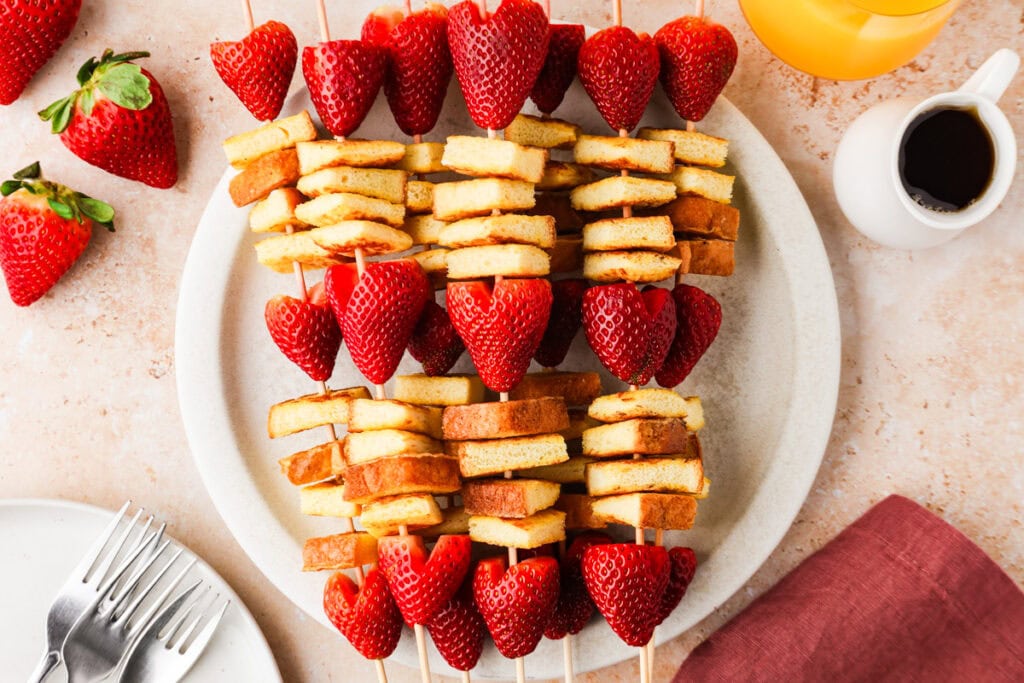 Skewers with alternating pieces of golden-brown French toast and halved strawberries are arranged on a white plate, with whole strawberries, a cup of syrup, orange juice, and forks nearby on a beige surface.