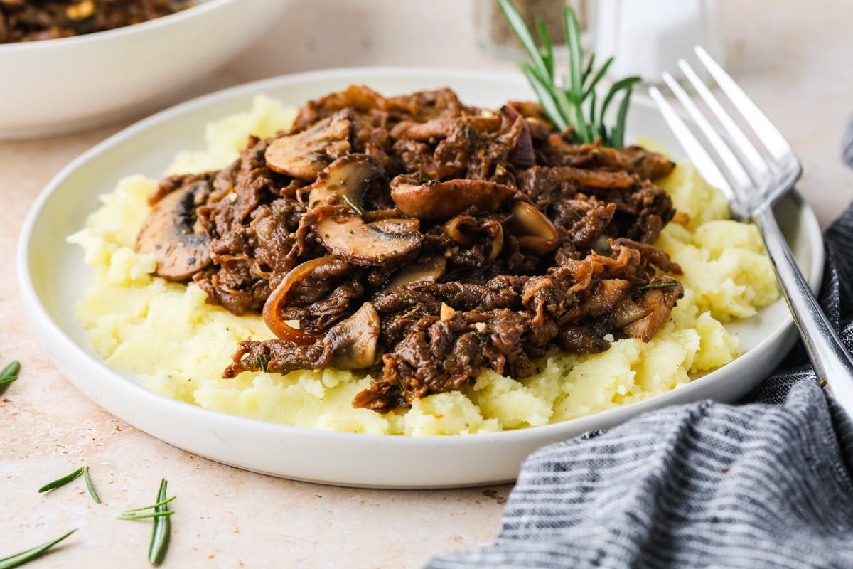 A plate of mashed potatoes topped with a savory mushroom and beef stew, inspired by a classic shaved steak recipe and garnished with fresh rosemary. A fork rests beside the plate on a light surface.
