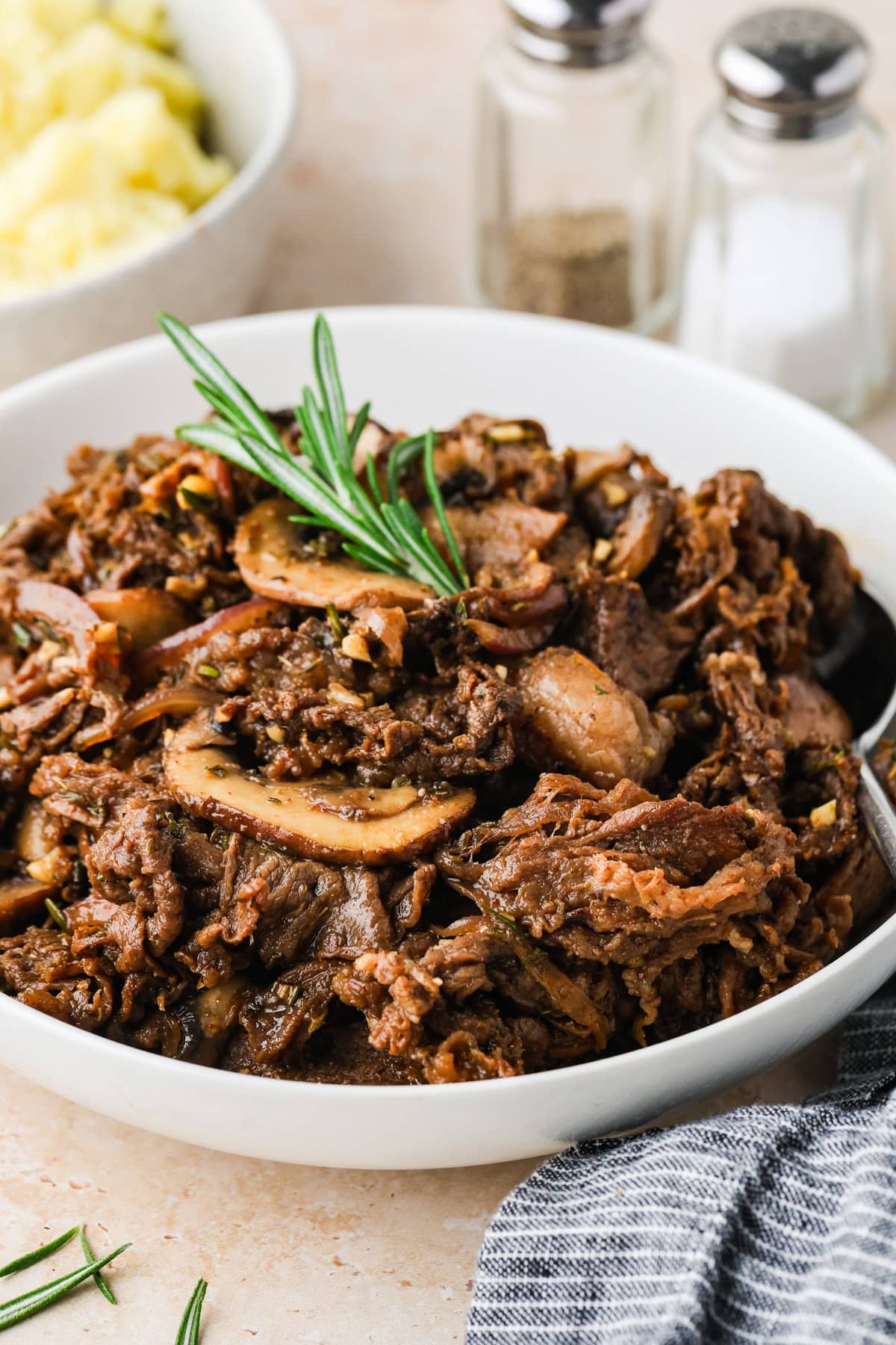 A white bowl filled with shredded beef and sliced mushrooms, inspired by a classic shaved steak recipe, garnished with a sprig of rosemary. Salt and pepper shakers and a bowl of mashed potatoes are blurred in the background.