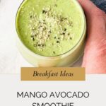 A hand holds a glass of mango avocado smoothie, topped with seeds. Text below reads: “Breakfast Ideas - Mango Avocado Smoothie. whensaltyandsweetunite.com.” A blue cloth is partially visible in the background.