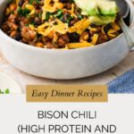 A bowl of bison chili topped with shredded cheddar cheese and avocado slices sits on a beige surface. Text below reads: “Easy Dinner Recipes: Bison Chili—High Protein, High Fiber, and packed with flavor.”.