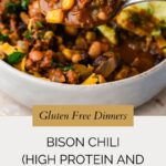 A spoonful of chunky bison chili with beans, corn, and herbs is held above a bowl. Text overlay reads: “Gluten Free Dinners – Bison Chili (High Protein and High Fiber).” Find this hearty bison chili recipe at whensaltyandsweetunite.com.