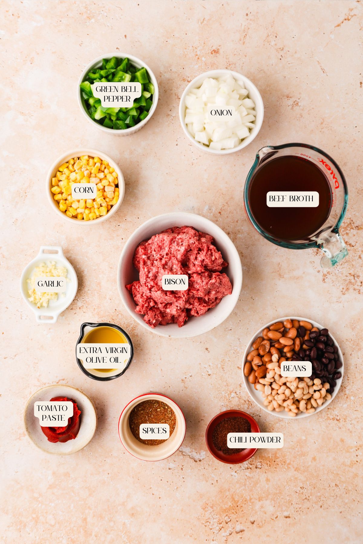Top-down view of bison chili ingredients in bowls on a light surface, including diced green bell pepper, onion, corn, garlic, olive oil, tomato paste, ground bison, beef broth, spices, chili powder, and beans. Labels identify each item.