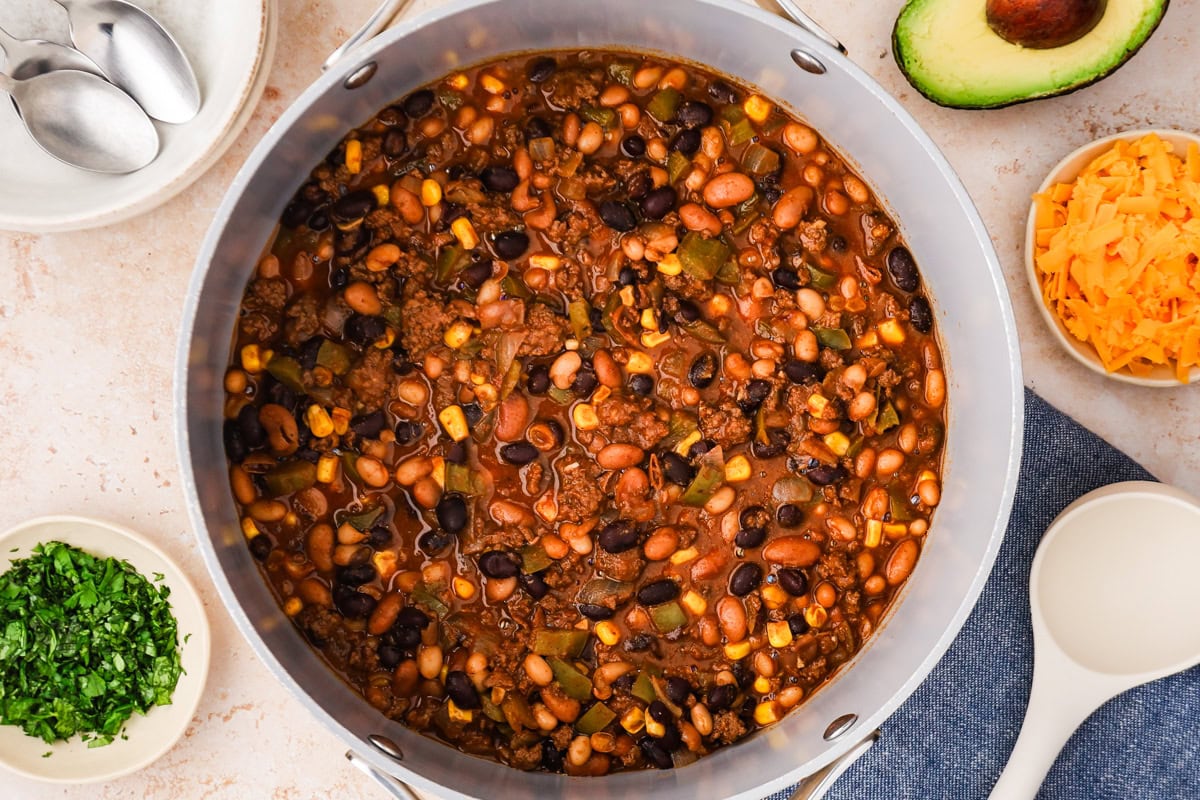 A large pot of bison chili with beans, corn, and ground meat sits on a counter, surrounded by bowls of chopped cilantro, shredded cheese, an avocado half, spoons, and a blue napkin with a white ladle.
