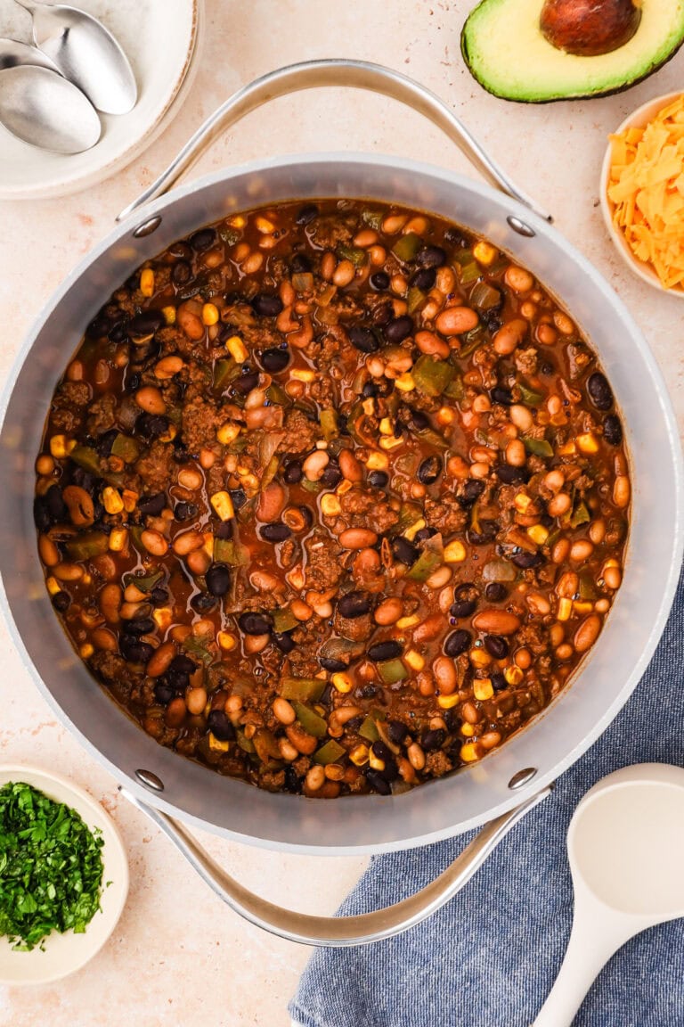 Overhead view of a large pot filled with hearty bison chili made from beans, corn, and ground meat. Surrounding the pot are bowls of shredded cheese, avocado, chopped herbs, and empty bowls with spoons.