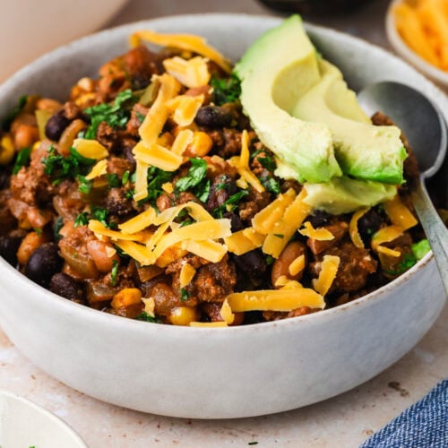 A bowl of bison chili topped with shredded cheddar cheese, sliced avocado, and chopped herbs, with a spoon inside. In the background are extra shredded cheese, chopped herbs, and half an avocado.