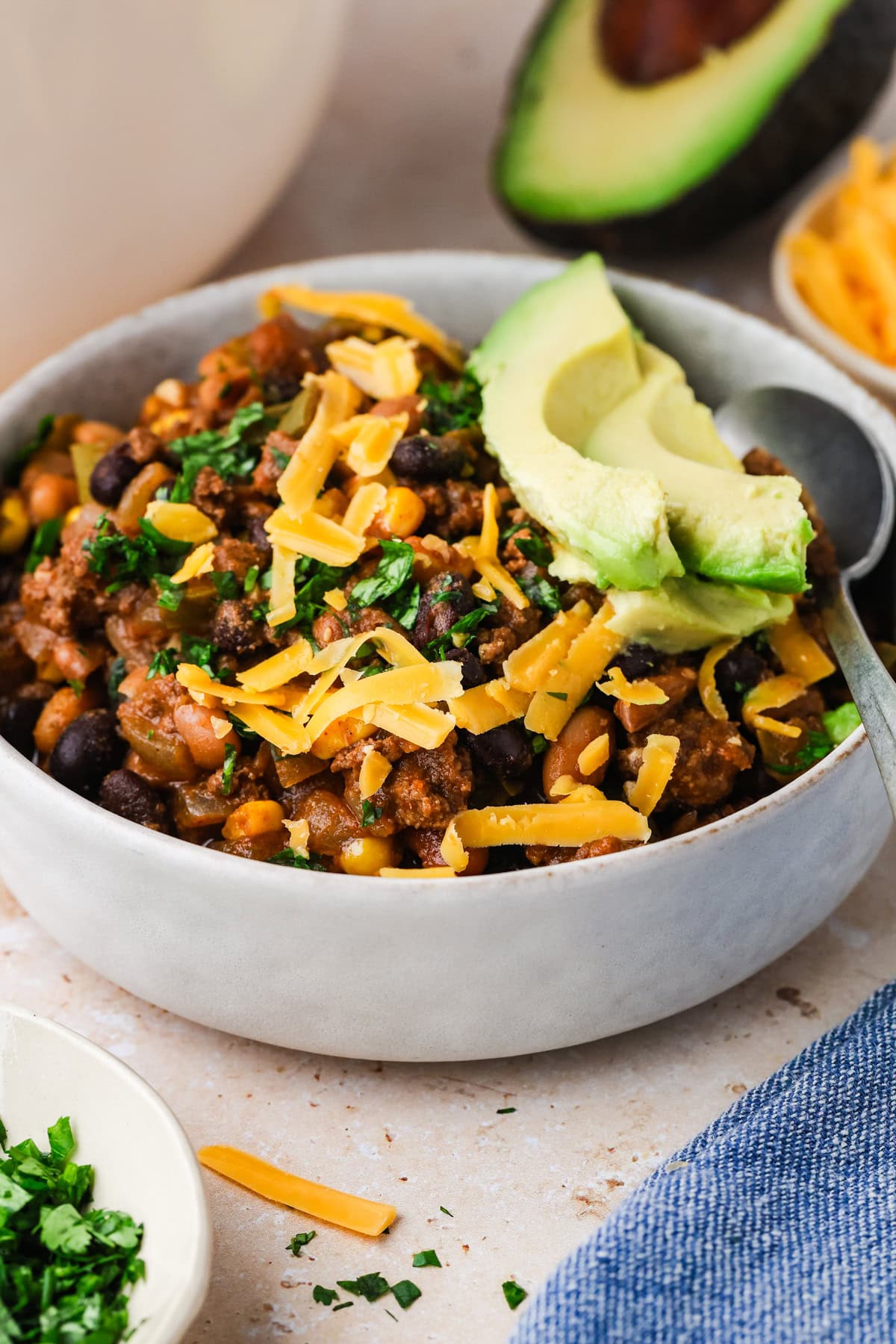 A bowl of bison chili topped with shredded cheddar cheese, sliced avocado, and chopped herbs, with a spoon inside. In the background are extra shredded cheese, chopped herbs, and half an avocado.