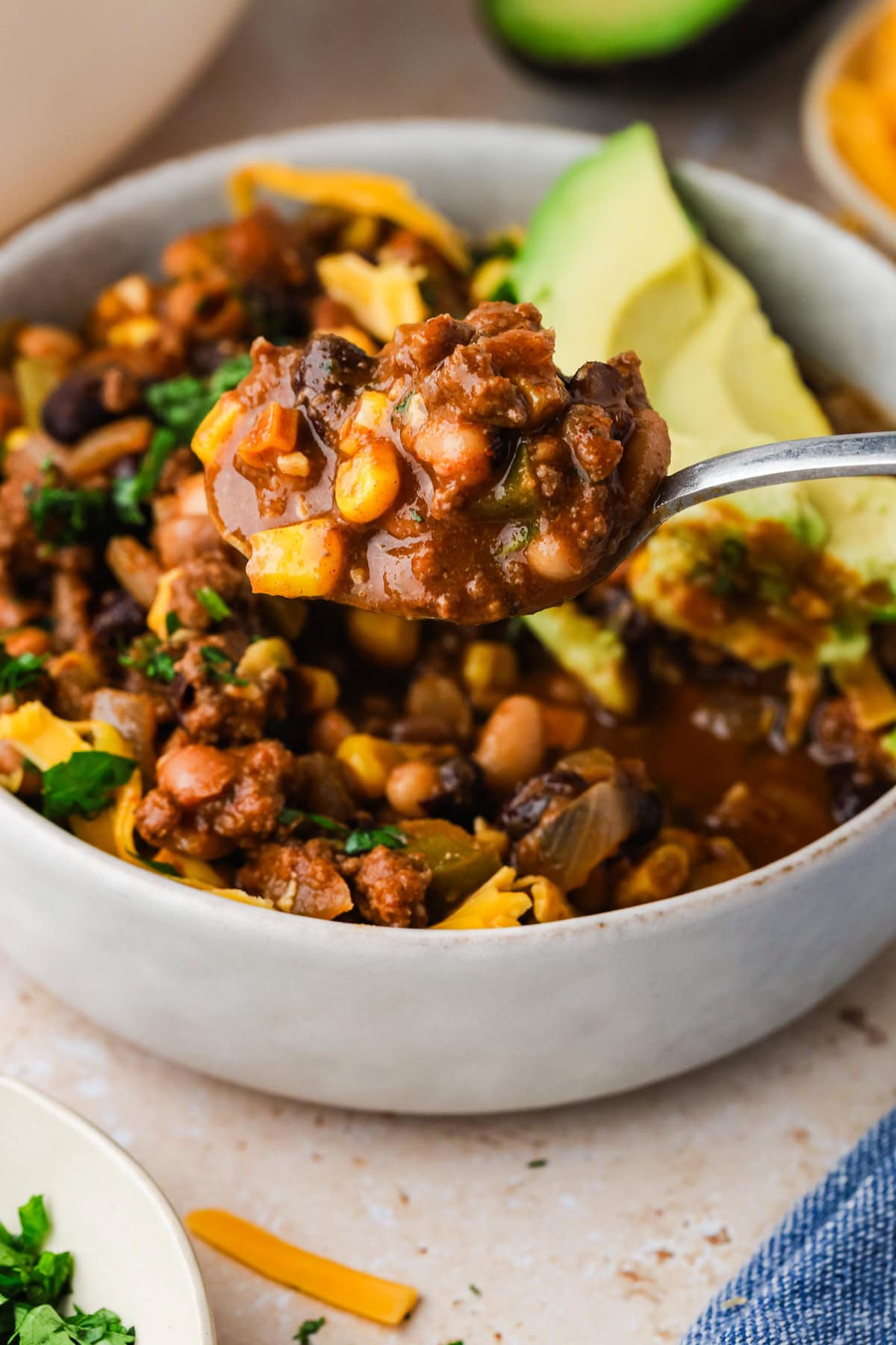 A close-up of a spoonful of bison chili with ground meat, beans, corn, and vegetables held above a bowl, garnished with sliced avocado and shredded cheese.