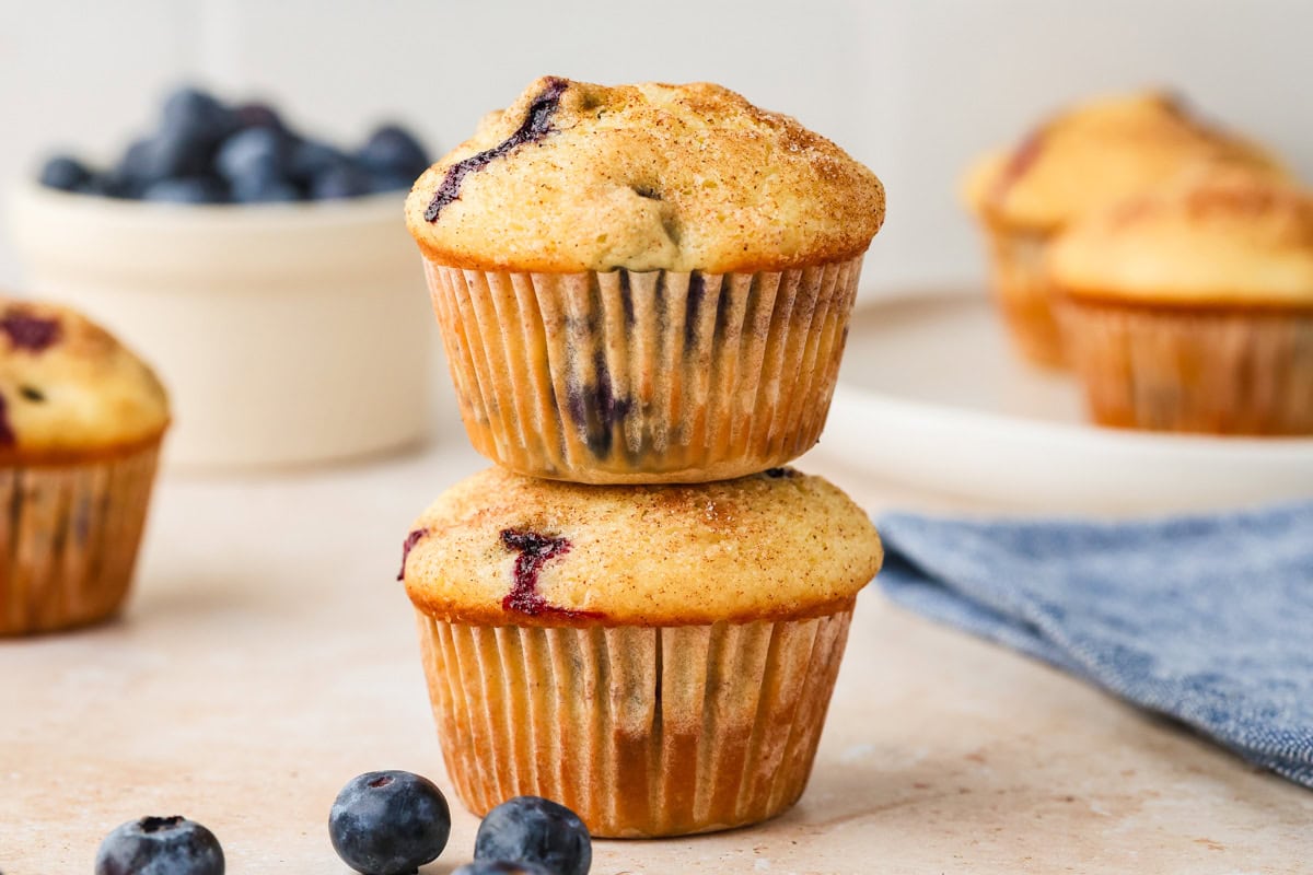 Two golden-brown Blueberry Buttermilk Muffins are stacked on a light surface, with fresh blueberries beside them. In the background, there’s a bowl of blueberries, more muffins, and a blue napkin.