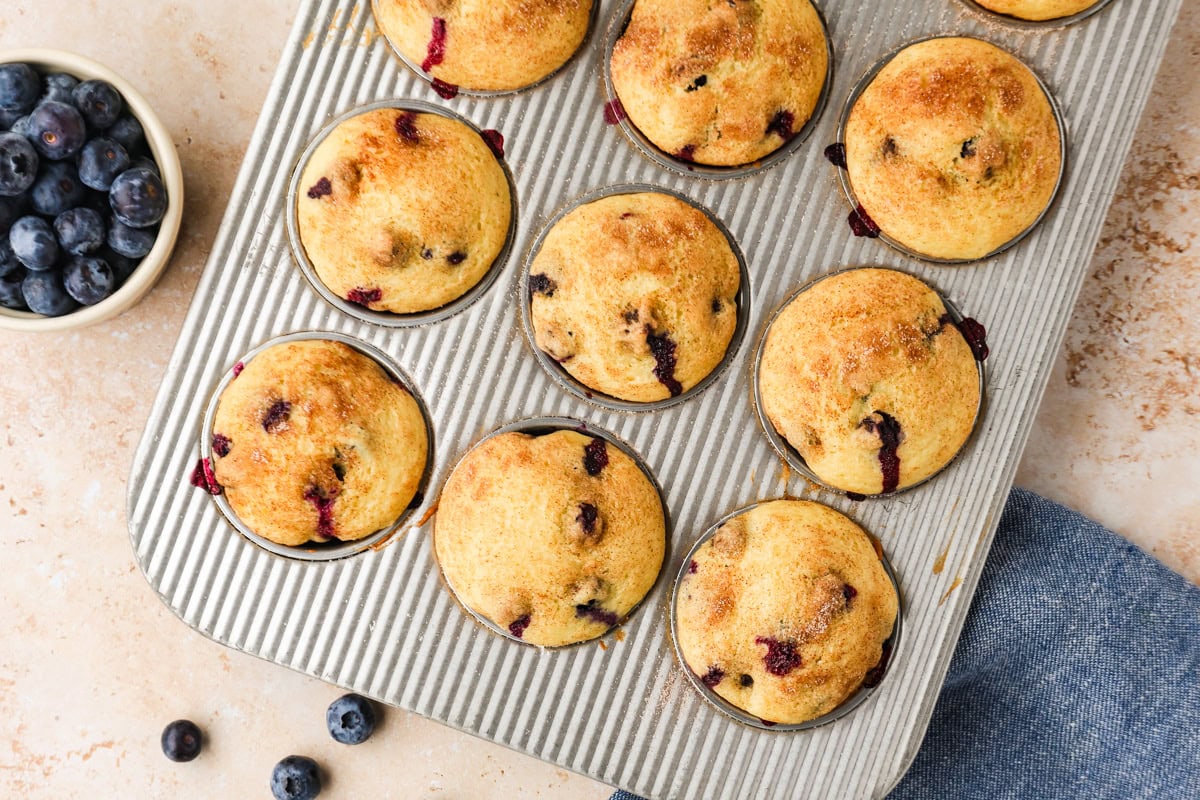 A muffin tin filled with freshly baked blueberry muffins sits on a light counter. A small bowl of blueberries and a few loose berries are nearby, with part of a blue napkin visible at the bottom right.
