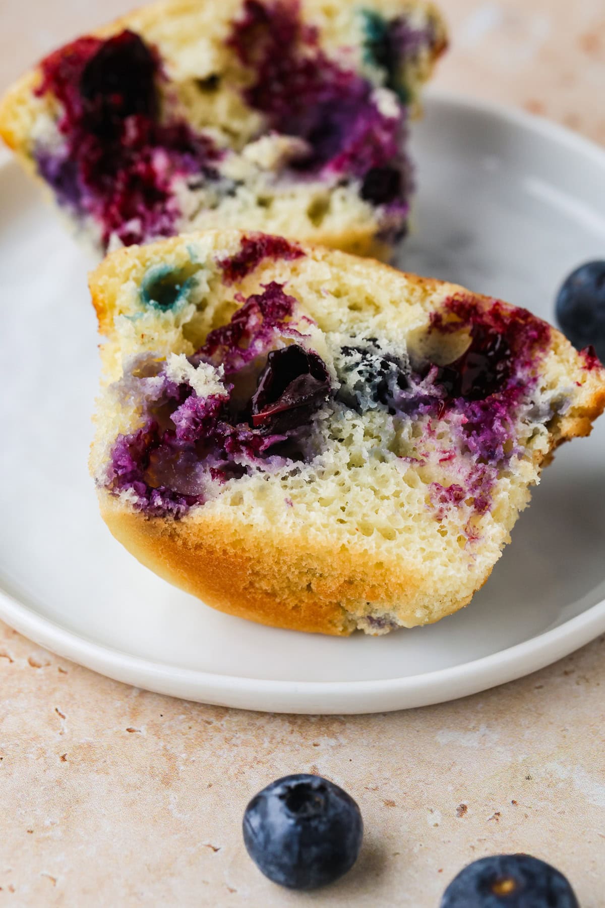 A close-up of a sliced Blueberry Buttermilk Muffin on a white plate, showcasing its soft, moist texture and vibrant blueberries inside. A few whole blueberries are scattered nearby on a light surface.