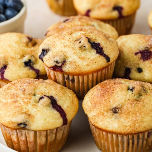 A plate of golden-brown Blueberry Buttermilk Muffins, some with visible blueberry pieces, sits on a light surface. A bowl of fresh blueberries is visible in the background.