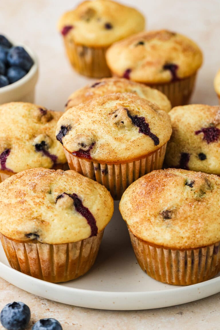 A plate of golden-brown Blueberry Buttermilk Muffins, some with visible blueberry pieces, sits on a light surface. A bowl of fresh blueberries is visible in the background.