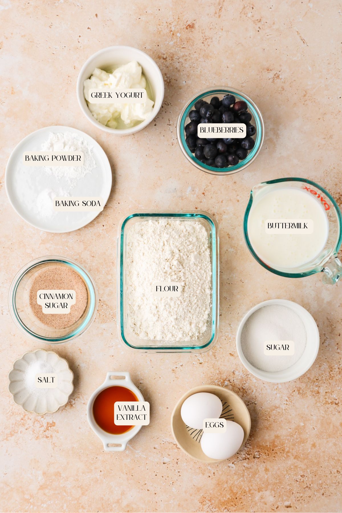 Top-down view of baking ingredients for Blueberry Buttermilk Muffins in bowls and containers, including flour, sugar, salt, cinnamon sugar, baking powder, Greek yogurt, blueberries, buttermilk, vanilla extract, and two eggs.