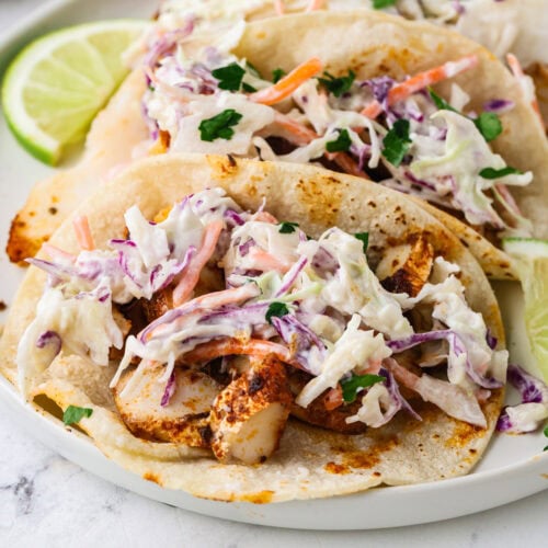 Three fish tacos topped with colorful coleslaw are served on a white plate, garnished with lime wedges. The tacos have grilled fish and a mix of shredded cabbage, carrots, and cilantro.