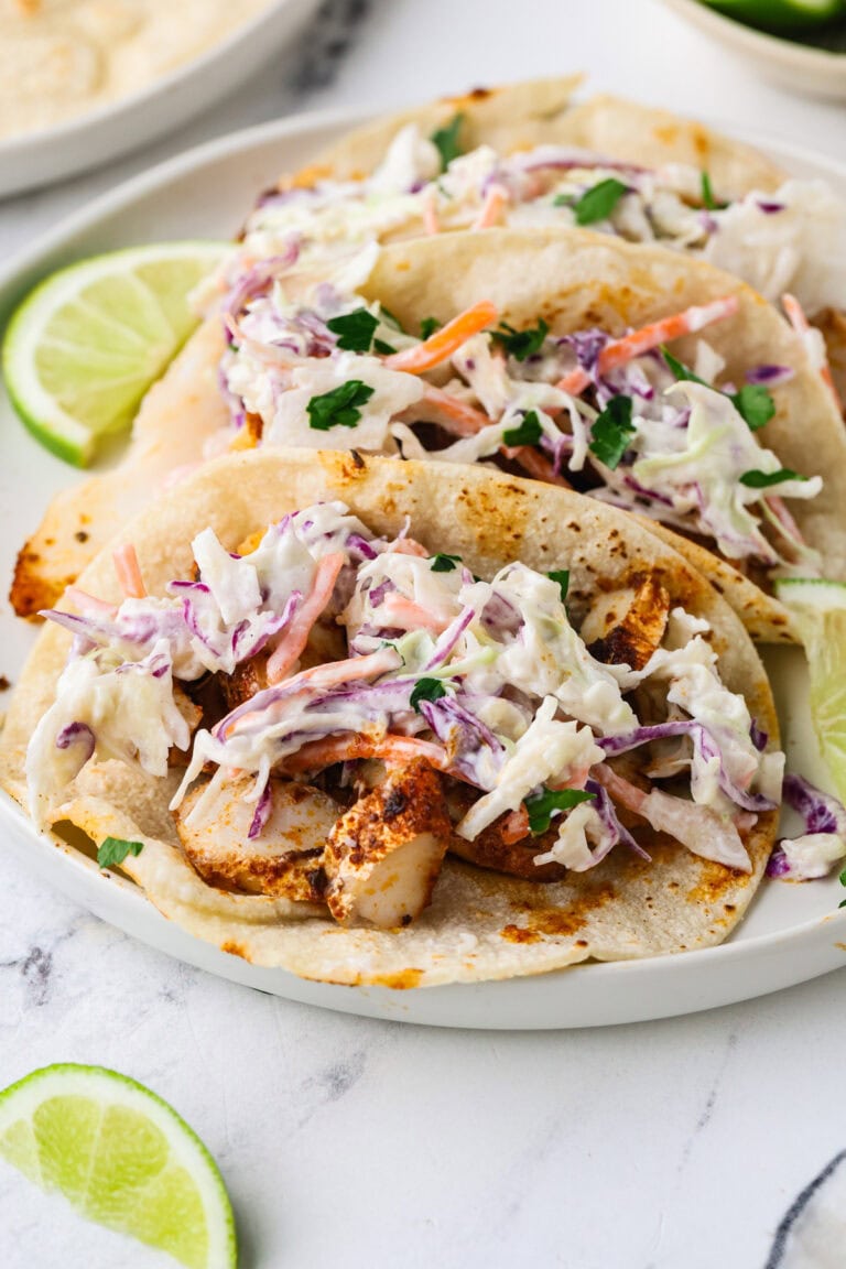 Three fish tacos topped with colorful coleslaw are served on a white plate, garnished with lime wedges. The tacos have grilled fish and a mix of shredded cabbage, carrots, and cilantro.