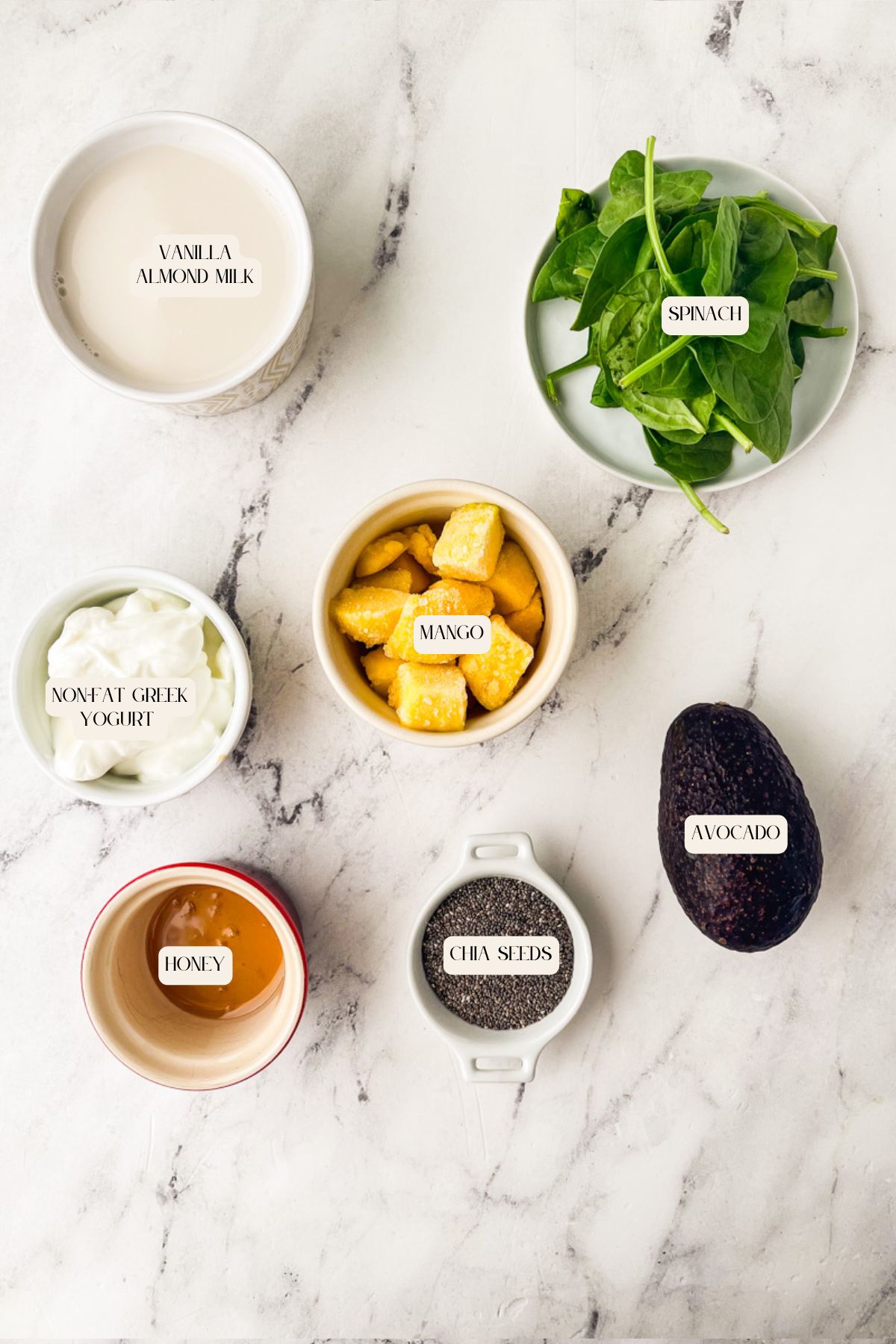 Six smoothie ingredients on a marble surface: a bowl of vanilla almond milk, a bowl of spinach leaves, a bowl of nonfat Greek yogurt, a bowl of mango pieces, a whole avocado, a dish of honey, and a dish of chia seeds.
