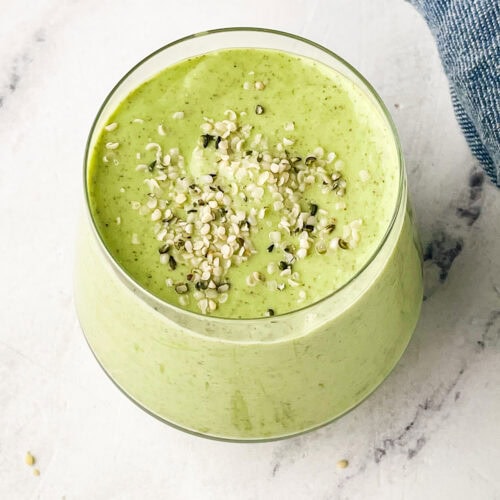 A glass filled with a creamy green mango avocado smoothie, topped with sprinkled hemp seeds, sits on a white marble surface next to a blue cloth.