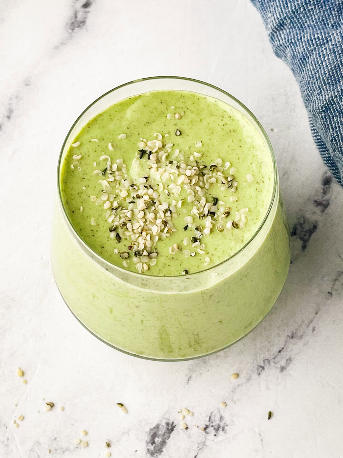 A glass filled with a creamy green mango avocado smoothie, topped with sprinkled hemp seeds, sits on a white marble surface next to a blue cloth.