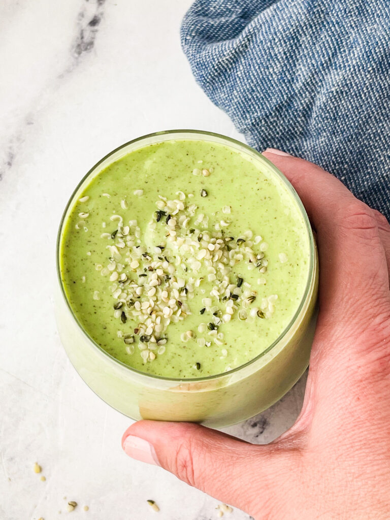 A hand holds a glass of mango avocado smoothie topped with sprinkled seeds, set on a white surface with a blue cloth partially visible in the background.