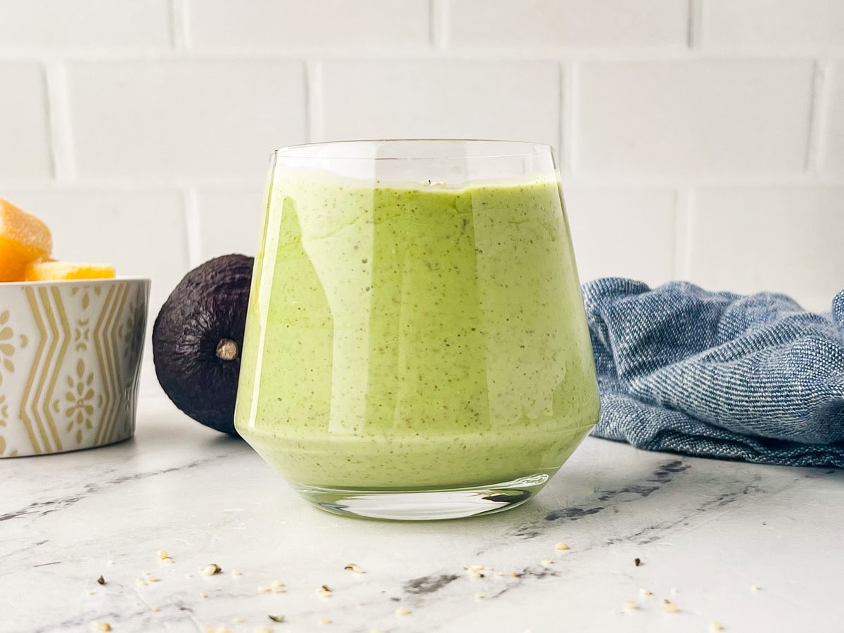 A glass filled with a creamy green smoothie sits on a white marble surface. In the background, there is an avocado, a bowl with fruit, and a blue cloth. The scene has a bright, fresh feel.