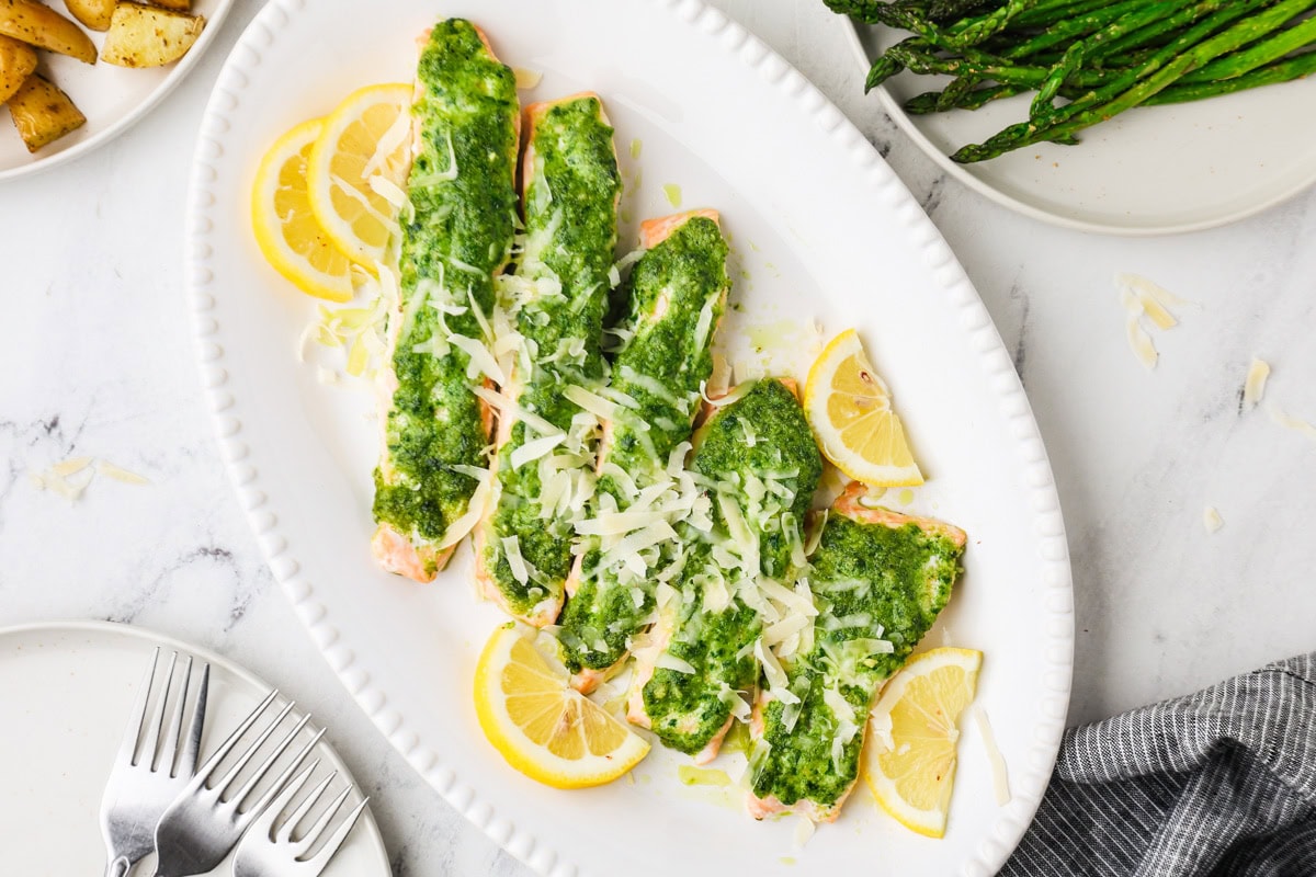 Three fillets of salmon topped with green pesto and shredded cheese are arranged on a white oval platter with lemon slices. Plates of roasted potatoes and asparagus are nearby on a white marble surface.