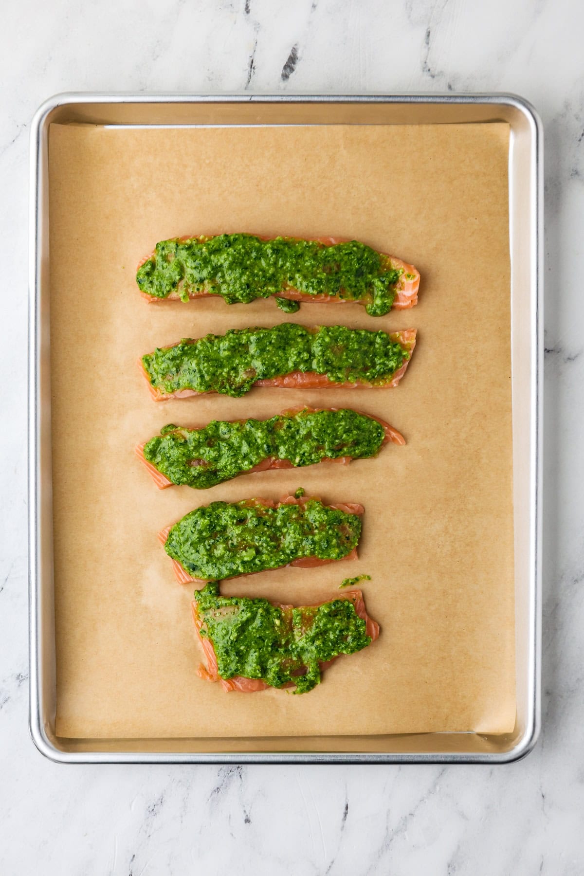 Five raw salmon fillets covered with green herb pesto are arranged in a row on a baking sheet lined with parchment paper, set on a white marble surface.
