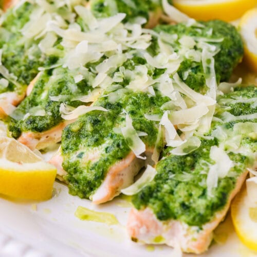 Salmon fillets topped with green pesto sauce and shredded cheese, garnished with lemon wedges, served on a white plate.