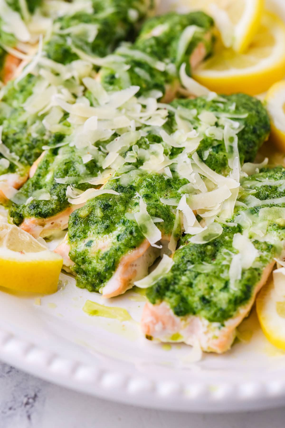 Salmon fillets topped with green pesto sauce and shredded cheese, garnished with lemon wedges, served on a white plate.