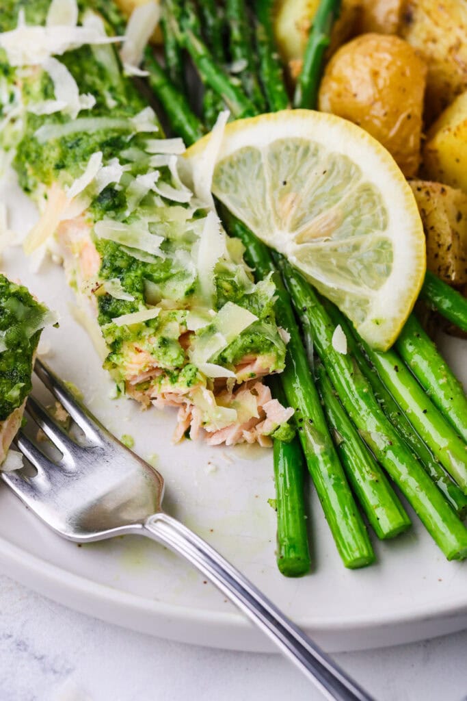 A close-up of a plate with herb-crusted salmon topped with shredded cheese, roasted asparagus, baby potatoes, and a slice of lemon. A fork rests beside the salmon on the white plate.