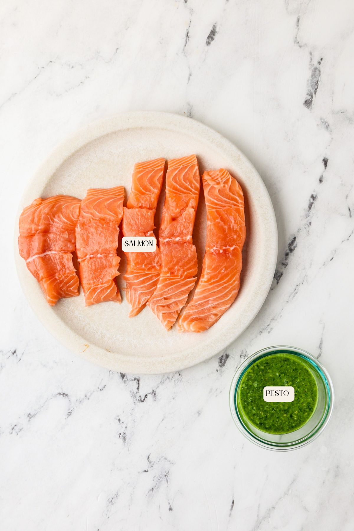 A white plate with four raw salmon fillets labeled SALMON and a small glass bowl with green pesto sauce labeled PESTO, both on a white marble surface.