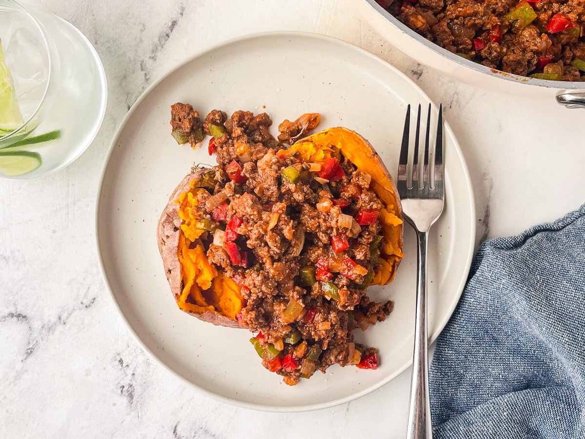 A baked sweet potato topped with seasoned ground beef, diced red and green peppers, and a hint of sugar free sloppy joe’s sauce sits on a white plate with a fork, next to a glass of water and a blue napkin.