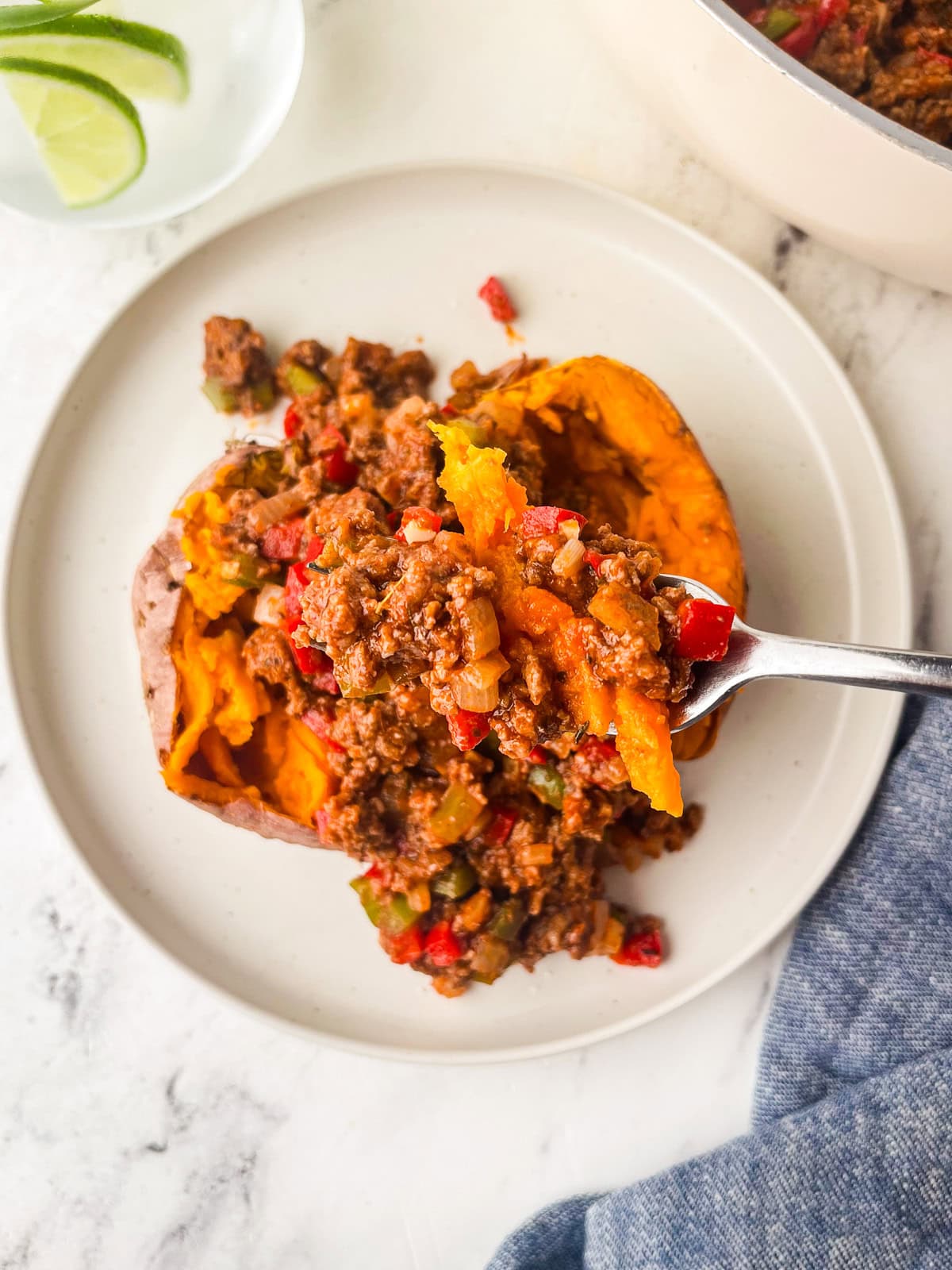 A baked sweet potato split open and topped with a savory sugar free sloppy joe's ground beef and vegetable mixture, served on a white plate with a glass of water and lime in the background.