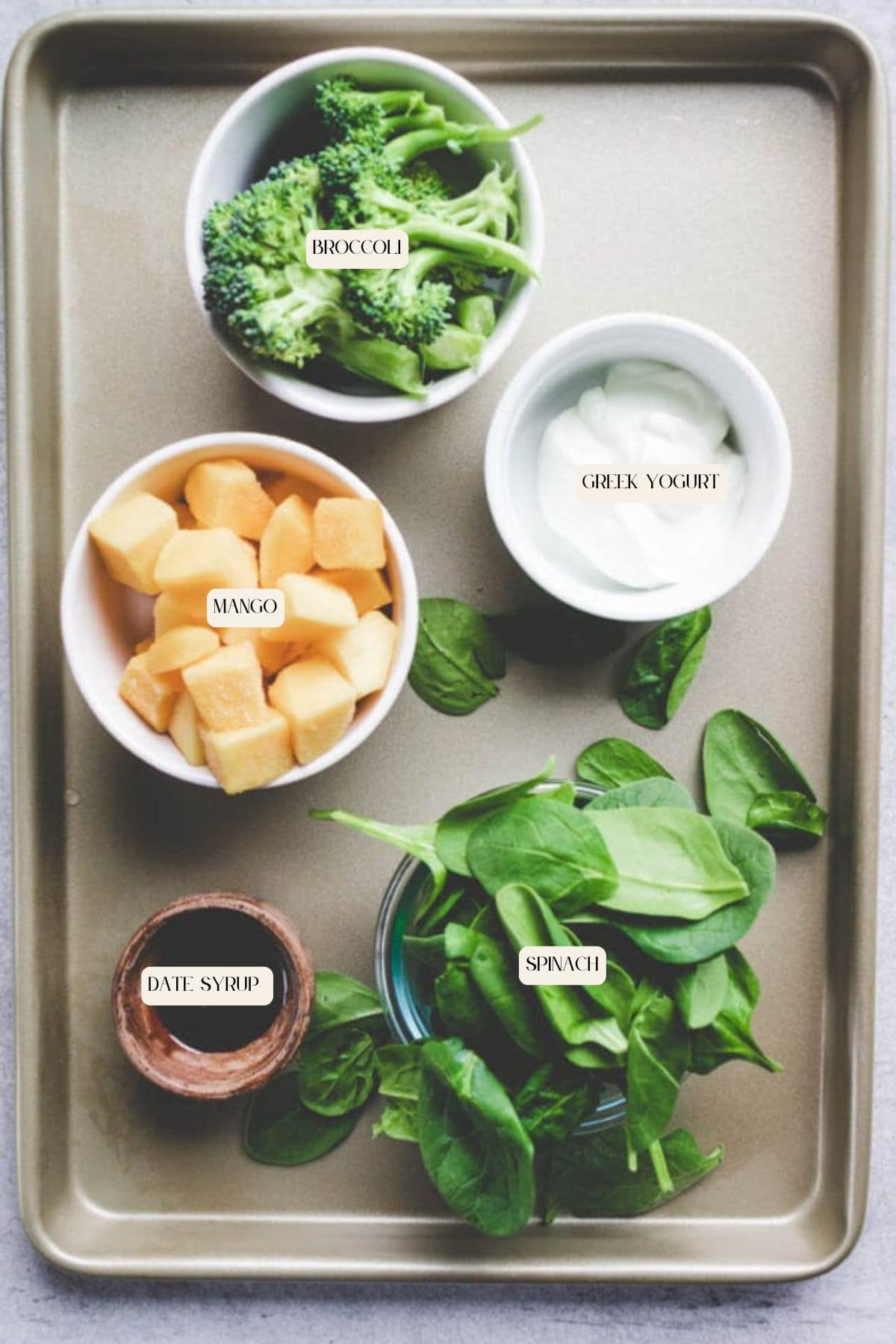A baking tray with bowls containing ingredients for a broccoli smoothie: broccoli, Greek yogurt, mango cubes, spinach leaves, and a small dish of date syrup, each labeled with text.
