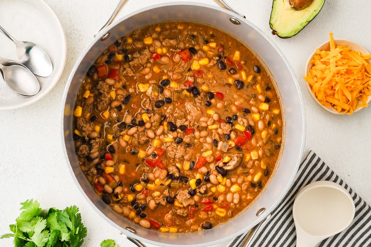 A pot of chili with ground meat, beans, corn, and red peppers on a white surface, surrounded by shredded cheese, half an avocado, fresh cilantro, and dining utensils.