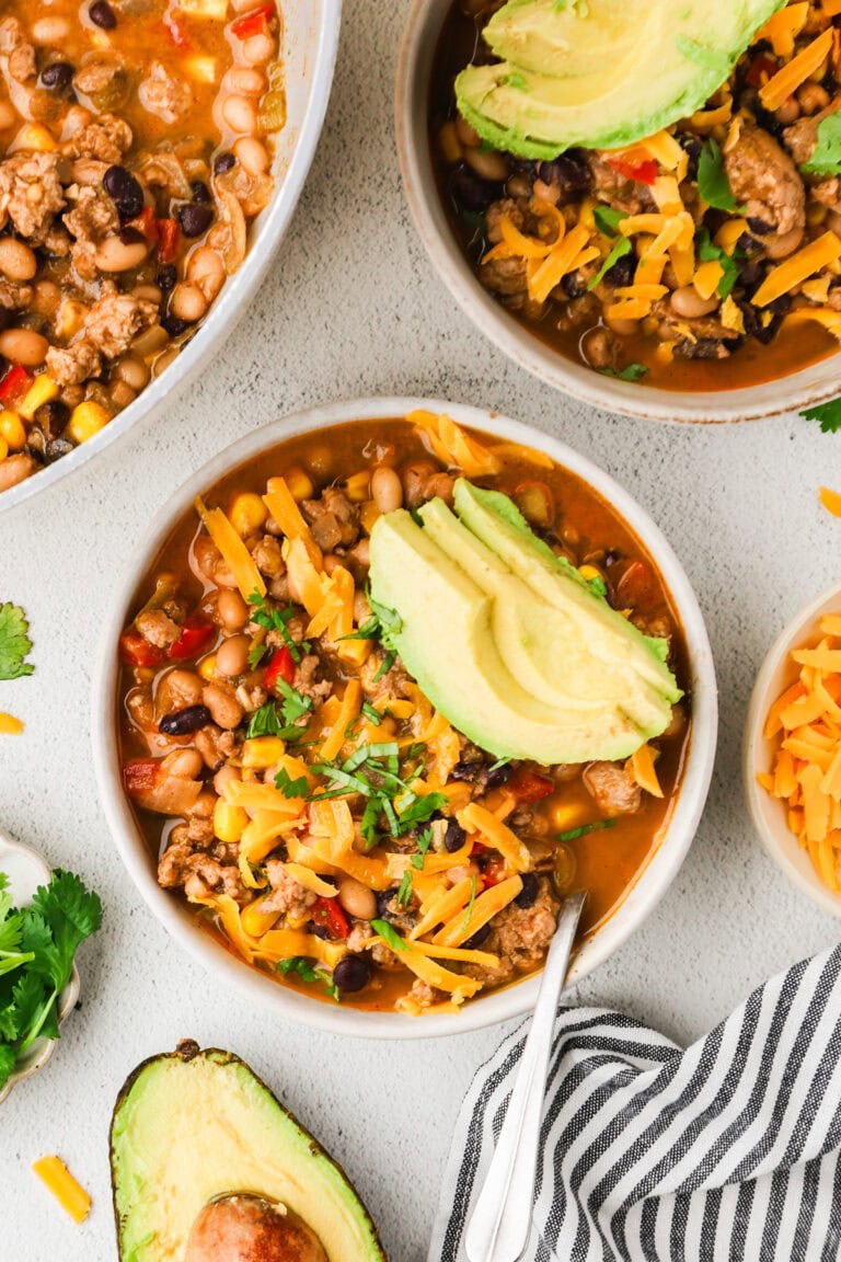 Two bowls of chili topped with shredded cheddar cheese, avocado slices, and cilantro. The chili contains ground meat, beans, corn, and peppers. Fresh avocado, shredded cheese, and cilantro garnish the table.