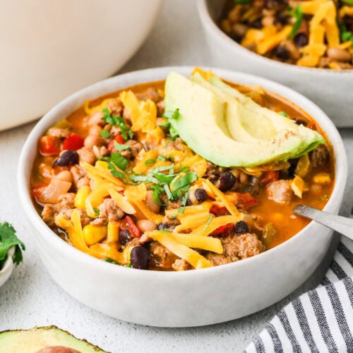 A bowl of chili topped with shredded cheese, chopped cilantro, and avocado slices, with another bowl, half an avocado, and a striped napkin nearby. The chili contains beans, ground meat, and vegetables.