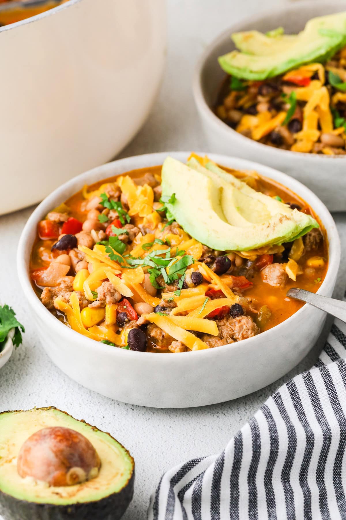 A bowl of chili topped with shredded cheese, chopped cilantro, and avocado slices, with another bowl, half an avocado, and a striped napkin nearby. The chili contains beans, ground meat, and vegetables.