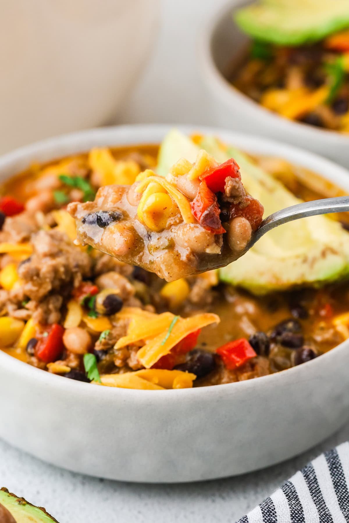 A close-up of a spoonful of taco soup with beans, corn, ground meat, diced tomatoes, red peppers, and melted cheese, held above a bowl of the same soup garnished with sliced avocado.