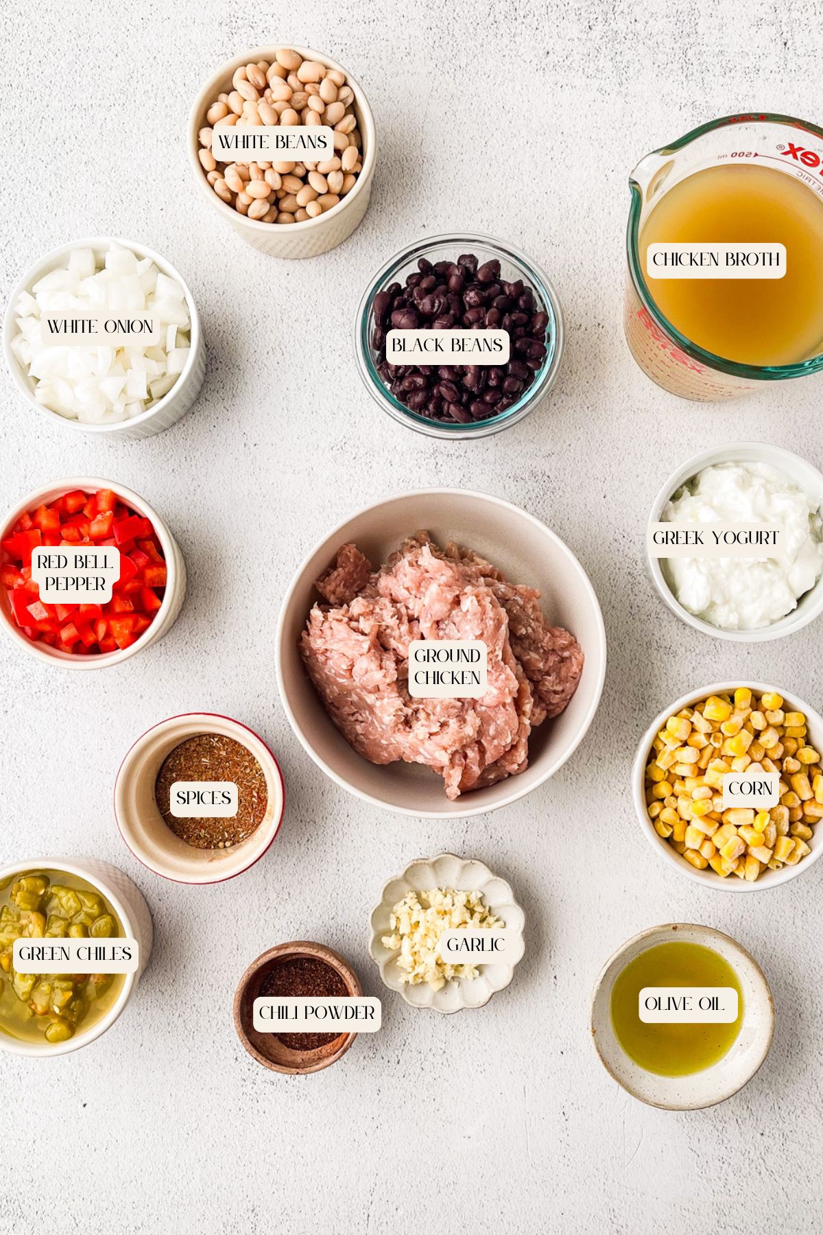 Ingredients for a chili recipe arranged on a white surface, including ground chicken, black and white beans, chicken broth, green chiles, corn, red bell pepper, white onion, garlic, Greek yogurt, olive oil, spices, and chili powder.