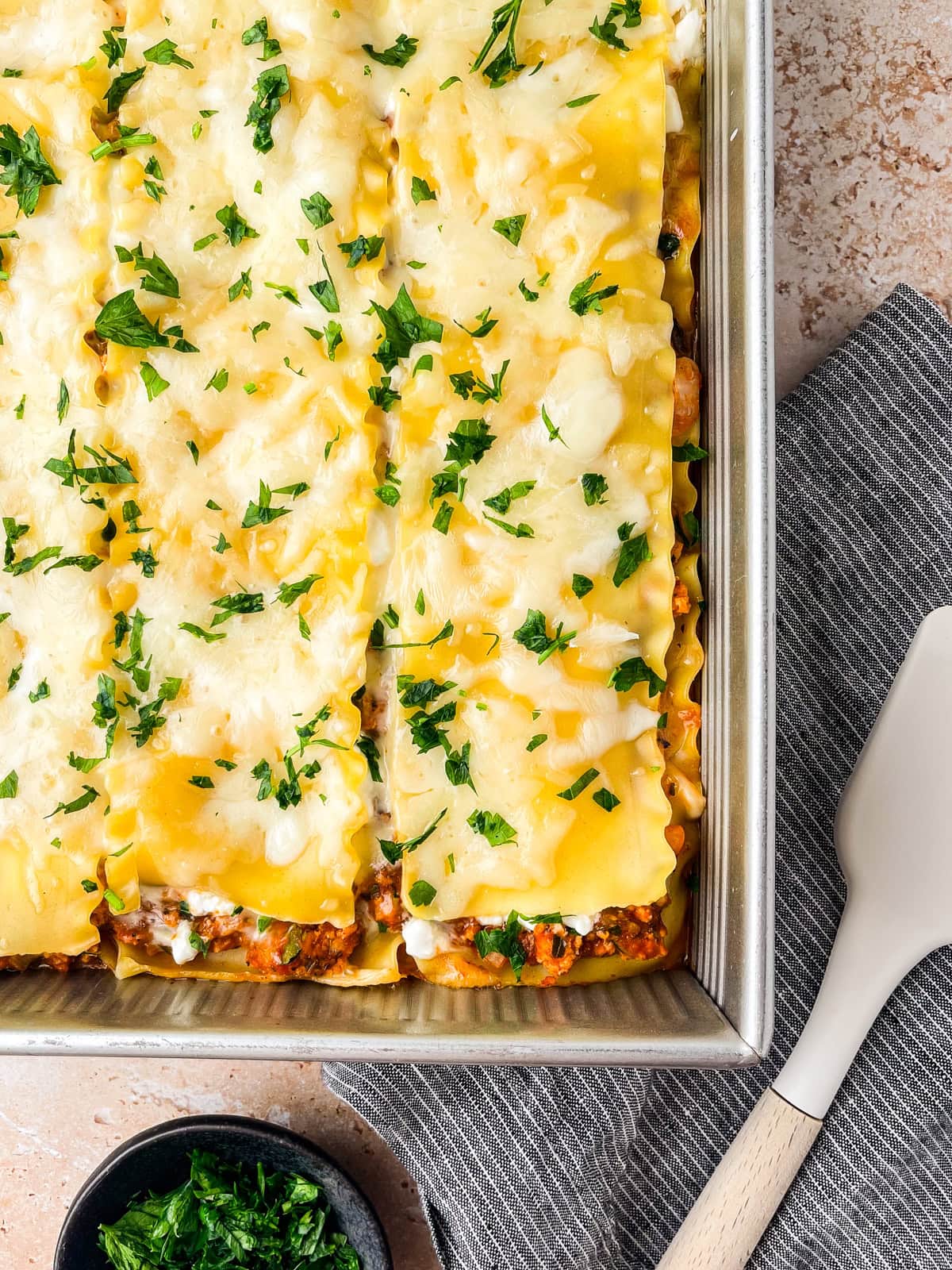 A baking dish filled with freshly baked Ground Chicken Lasagna, topped with melted cheese and chopped parsley. A spatula and a small bowl of extra parsley are placed next to the dish on a gray striped cloth.