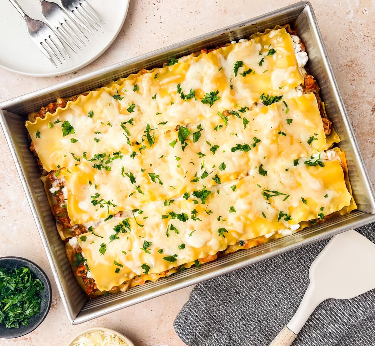 A rectangular baking dish filled with baked Ground Chicken Lasagna, topped with melted cheese and chopped parsley. Plates, forks, a bowl of herbs, and a spatula are arranged nearby on a light surface.