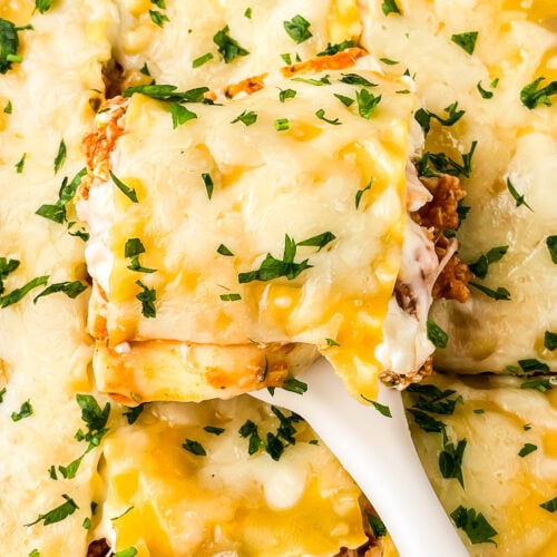 A close-up of a cheesy Ground Chicken Lasagna topped with chopped parsley. A spatula lifts out a square piece, revealing layers of pasta, savory meat sauce, and melted cheese.