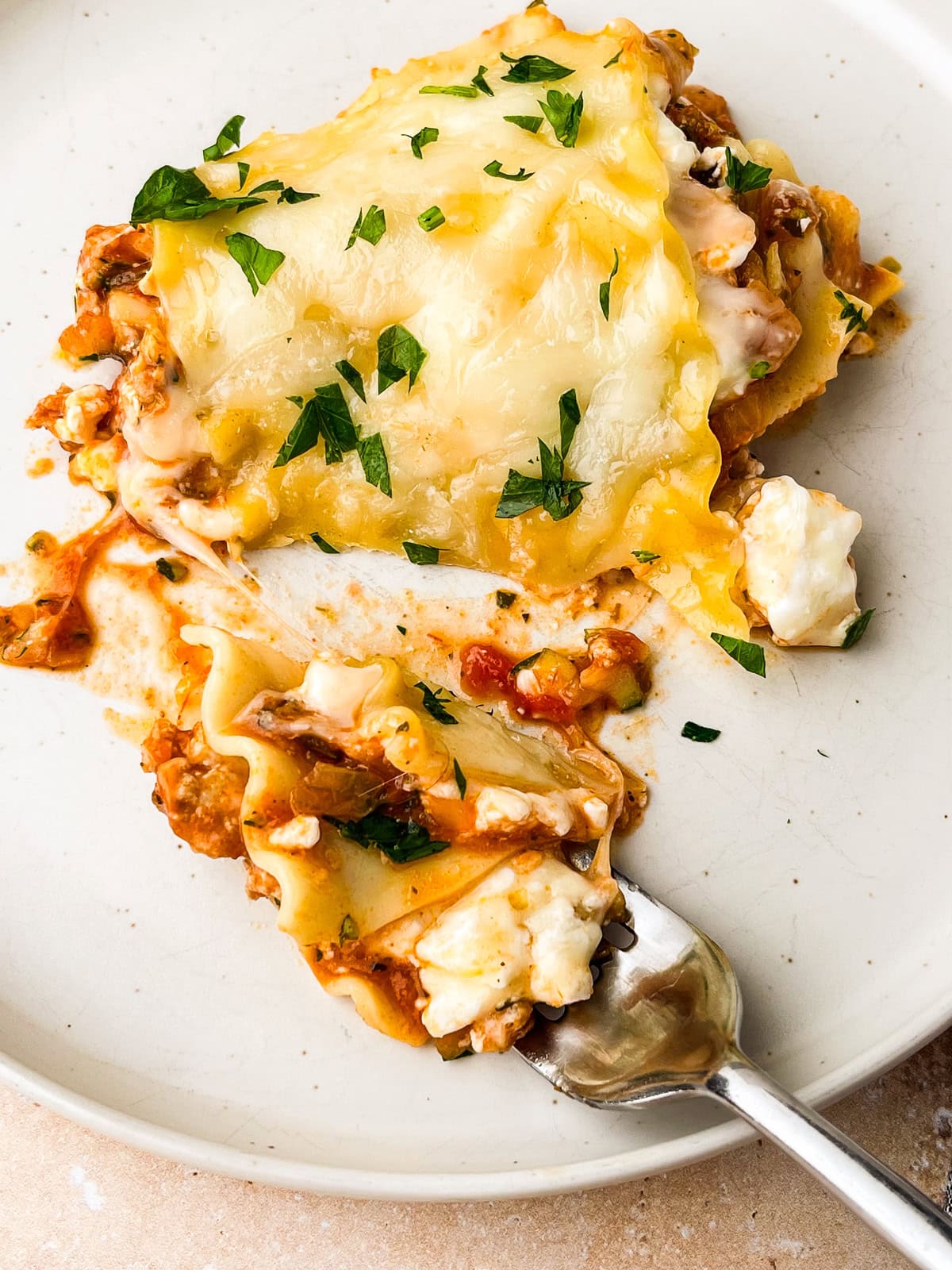 A slice of Ground Chicken Lasagna with melted cheese and parsley on top sits on a white plate. A fork holds a bite showing layers of pasta, tomato sauce, ground chicken, and creamy ricotta cheese.