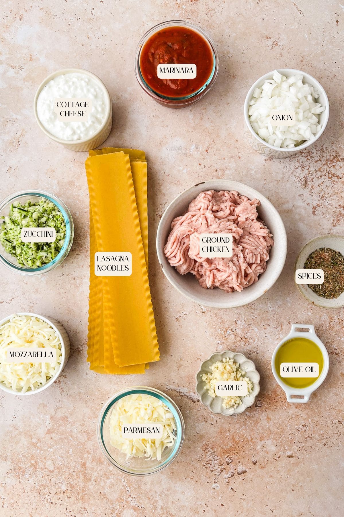 Top-down view of Ground Chicken Lasagna ingredients, featuring cottage cheese, marinara sauce, chopped onion, ground chicken, dry spices, olive oil, garlic, parmesan, mozzarella, shredded zucchini, and uncooked lasagna noodles.
