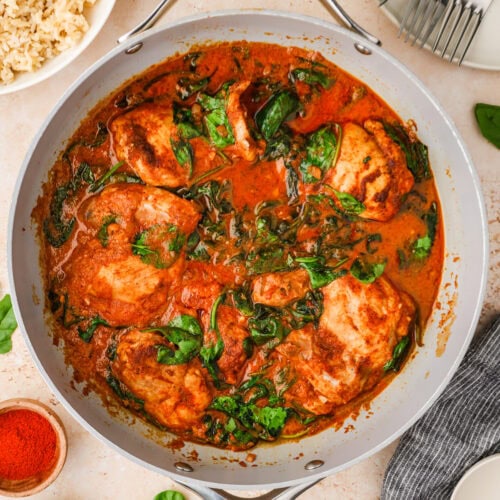 A skillet filled with chicken breasts cooked in a rich tomato-based sauce with wilted spinach. Nearby are bowls of rice, plates, forks, spices, and scattered fresh spinach leaves on a light surface.