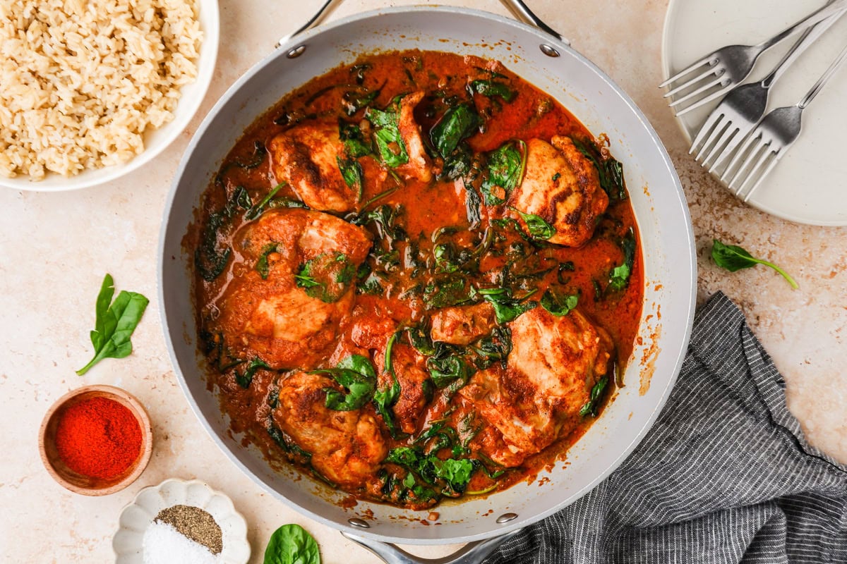 A skillet filled with chicken breasts cooked in a tomato-based sauce with wilted spinach. Surrounding the skillet are a bowl of brown rice, plates with forks, a striped napkin, and small dishes of spices.