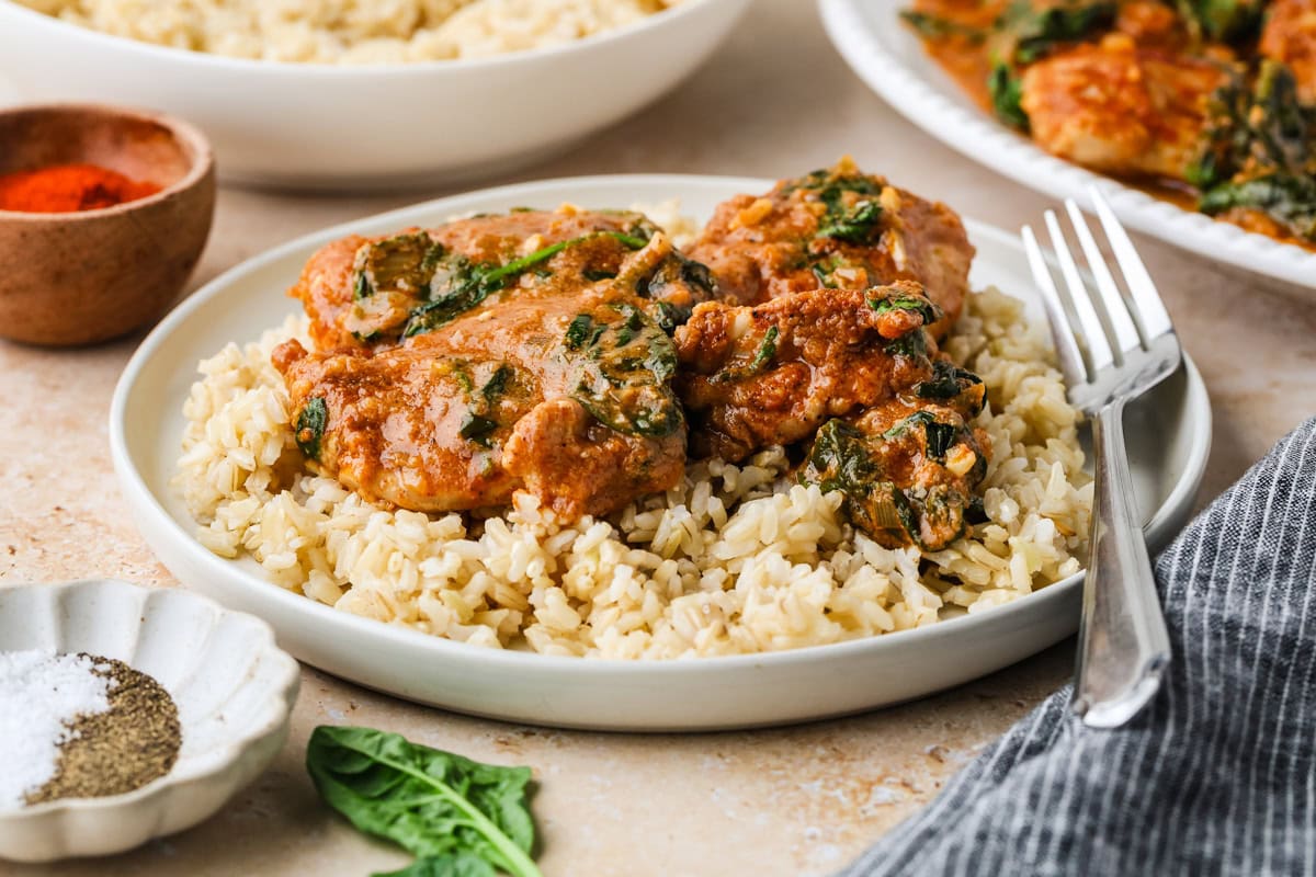 A white plate filled with brown rice topped with saucy chicken pieces and wilted spinach, with a fork beside it. Nearby are bowls of pepper, salt, and a dish with more of the chicken and sauce.